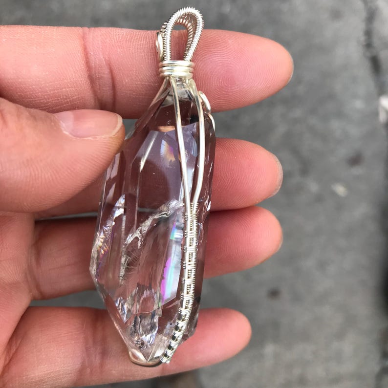 May include: A clear, elongated quartz crystal pendant encased in a silver-colored wire wrap. The crystal features internal fractures that create rainbow light refractions, and the wire forms a secure loop at the top for hanging as a necklace.