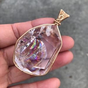 May include: A clear, faceted crystal pendant encased in a gold-colored wire wrap. The crystal features internal fractures that create a vibrant, multi-colored rainbow reflection within the stone. A triangular wire bail sits at the top of the pendant.