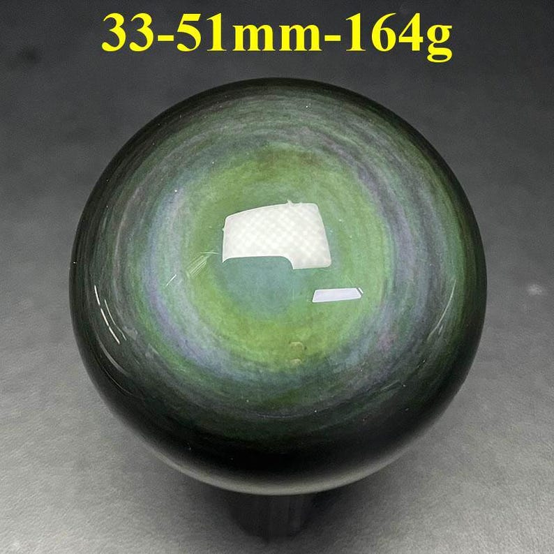 May include: A polished, spherical obsidian stone with a swirling pattern of green, gray, and black. The text "33-51mm-164g" is visible at the top. The stone measures approximately 1.3 to 2 inches in diameter and weighs about 5.8 ounces.