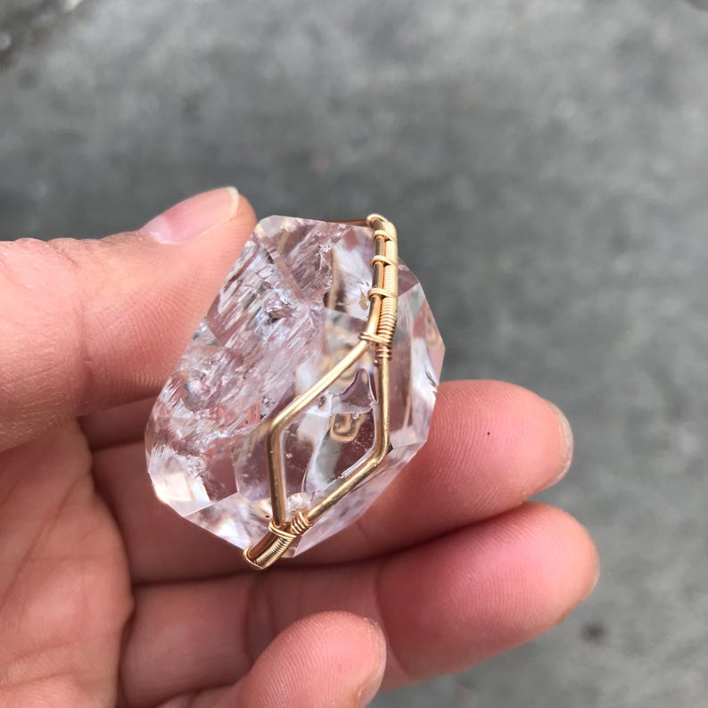May include: A clear, faceted quartz crystal pendant secured with a gold-colored wire wrap. The wire is coiled tightly around the edges of the stone, creating a geometric frame that holds the crystal in place. The stone has visible internal fractures.