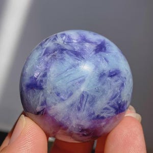 May include: A polished sphere of fluorite, showcasing a blend of light blue, white, and deep purple hues. The crystal ball has a smooth, rounded shape, with a translucent quality. The colors are swirled and layered, creating a unique pattern.
