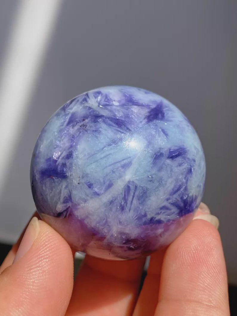 May include: A polished sphere of fluorite, showcasing a blend of light blue, white, and deep purple hues. The crystal ball has a smooth, rounded shape, with a translucent quality. The colors are swirled and layered, creating a unique pattern.