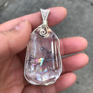 May include: A clear, faceted quartz crystal pendant encased in a silver-colored wire wrap. The crystal features internal fractures and a visible rainbow-colored light refraction within its structure. The wire forms a decorative bail at the top.