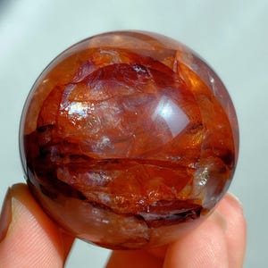 Puede incluir: Una bola de cristal esférica con un interior marrón rojizo. La superficie pulida refleja la luz, creando reflejos y sombras. El cristal parece translúcido, revelando patrones y texturas internas. Objeto decorativo.