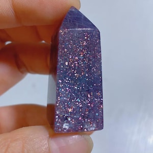 Pu&ograve; includere: Una torre di cristallo viola lucido con punta esagonale, caratterizzata da una base traslucida incastonata con numerose particelle di glitter riflettenti multicolori che brillano in tonalit&agrave; rosa, blu e argento sulla superficie della pietra.