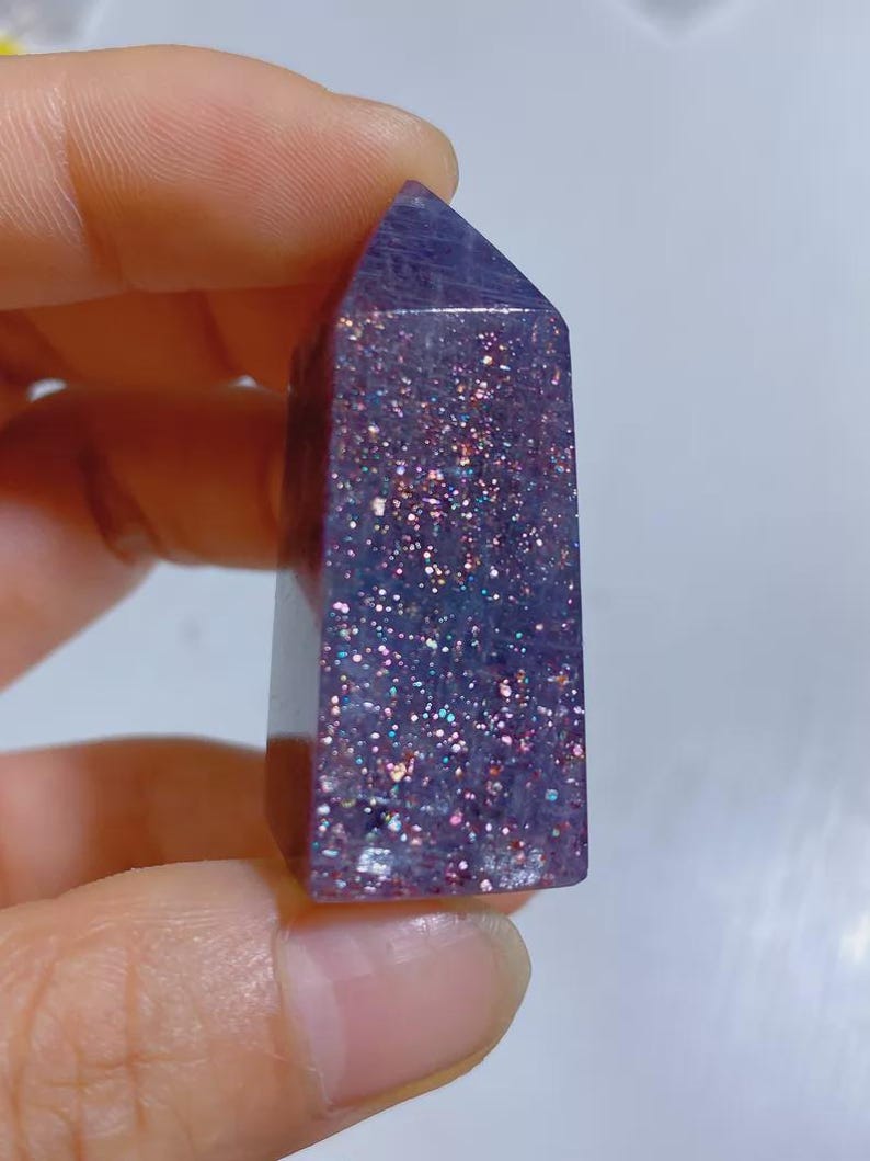 Pu&ograve; includere: Una torre di cristallo viola lucido con punta esagonale, caratterizzata da una base traslucida incastonata con numerose particelle di glitter riflettenti multicolori che brillano in tonalit&agrave; rosa, blu e argento sulla superficie della pietra.