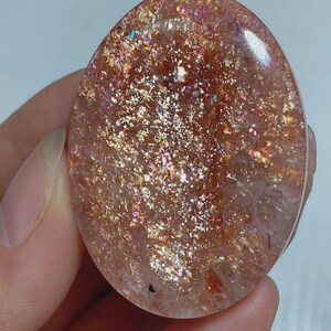 Puede incluir: Un cabujón de piedra de sol de forma ovalada con una base translúcida en tonos melocotón, lleno de densas inclusiones metálicas brillantes de color cobre y oro que crean un efecto reflectante y centelleante en toda la superficie pulida.