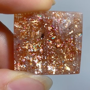 Op de afbeelding: Een vierkante zonnesteen cabochon met een doorschijnende perzikkleurige basis en dichte, reflecterende metallic koperen en gouden insluitingen die een glinsterend, iriserend effect creëren over het gehele oppervlak van de edelsteen.