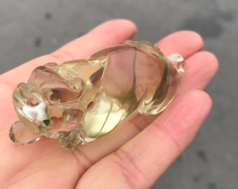 Op de afbeelding: Een helder, gebeeldhouwd kristallen beeldje van een klein dier, mogelijk een beer of een hond, met een lichtgele tint. Het beeldje wordt in een hand gehouden, wat het gedetailleerde vakmanschap en de doorschijnende kwaliteit laat zien. Het dier ligt.