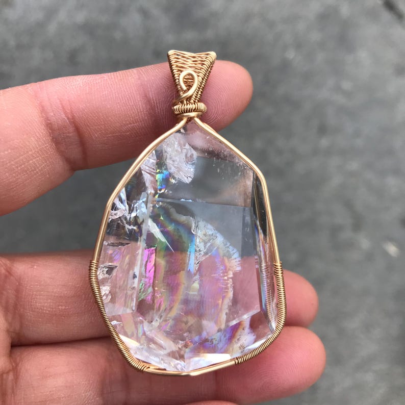 May include: A clear, irregularly shaped quartz crystal pendant wrapped in gold-colored wire. The crystal features internal fractures that create a rainbow-like iridescent effect. The wire forms a decorative bail at the top for suspension.