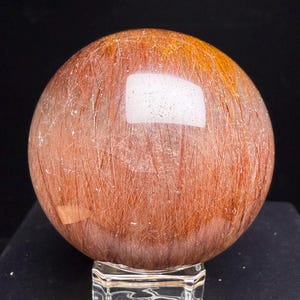 Peut inclure: Une sphère en quartz rutile poli présentant une base transparente remplie d'inclusions denses en forme d'aiguilles dans des tons brun rougeâtre et cuivre. La sphère affiche une surface lisse et réfléchissante avec des motifs minéraux naturels.