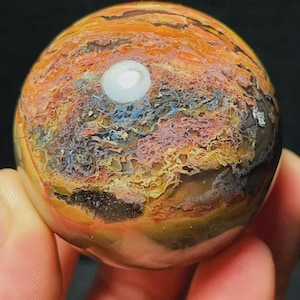 Puede incluir: Una piedra esférica pulida que presenta un patrón complejo de colores arremolinados, incluyendo naranja intenso, amarillo dorado, rosa suave y marrón oscuro. La superficie muestra inclusiones intrincadas tipo musgo y un reflejo blanco.