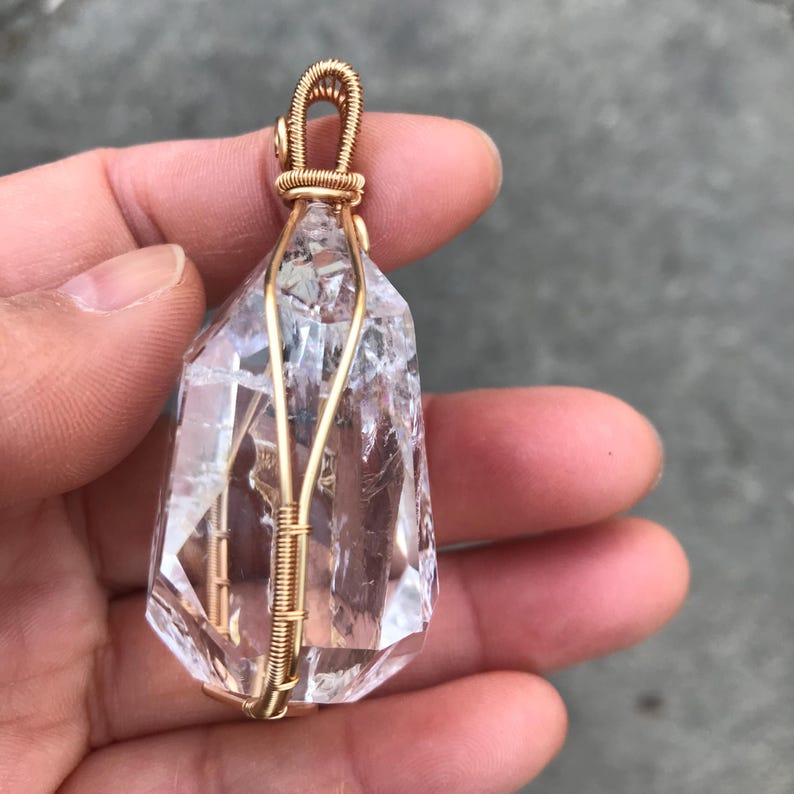 May include: A clear, faceted quartz crystal pendant encased in a gold-colored wire wrap. The wire forms a secure cage around the stone and extends upward to create a small, coiled loop at the top for hanging, showcasing the stone's internal texture.