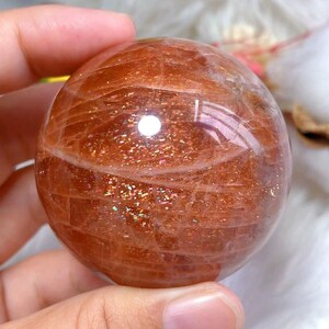 Peut inclure: Une pierre de soleil polie en forme de sphère, présentant une teinte orange translucide et chaude avec des fractures internes et des inclusions scintillantes semblables à des paillettes métalliques qui captent la lumière sur sa surface lisse.