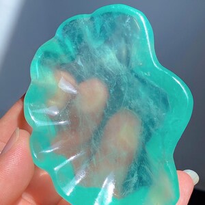Op de afbeelding: Een doorschijnend, turquoise-kleurig, bloemvormig kristal. Het kristal wordt in een hand gehouden en het licht schijnt erdoorheen, waardoor een gloeiend effect ontstaat. De randen van het kristal zijn glad en afgerond. Het is een uniek decoratief item.