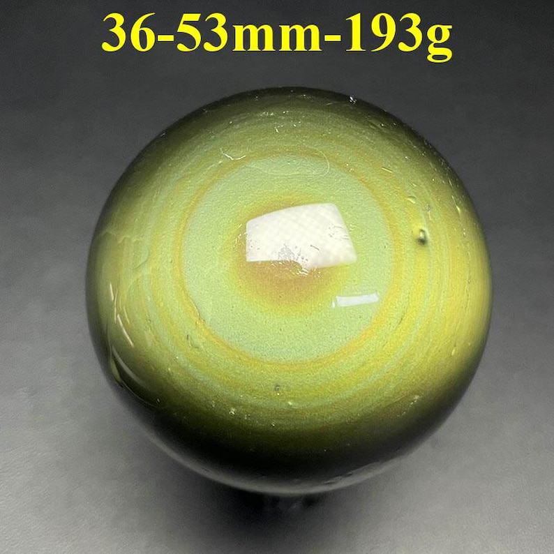 May include: A polished, spherical gemstone with concentric bands of green and yellow hues. The stone has a weight of 193g and a diameter between 36-53mm. A white, rectangular inclusion is visible on the surface.