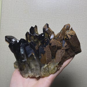 Pode incluir: Um aglomerado de cristais de quartzo fumê com pontas escuras e translúcidas e depósitos minerais castanhos, opacos e texturizados na base e nas laterais. A formação consiste em múltiplas pontas de cristal verticais e irregulares com níveis variados.