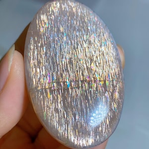 Op de afbeelding: Een ovaalvormige zonnesteen cabochon met een doorschijnende, licht perzikkleurige basis en opvallende, glinsterende metalen insluitingen die een levendig, veelkleurig aventurescentie-effect creëren op het gepolijste, gladde oppervlak.