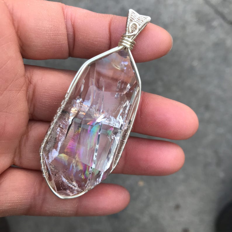 May include: A clear, elongated, faceted crystal pendant encased in a silver-colored wire-wrapped frame. The crystal features internal fractures that create a rainbow-like iridescent effect when light hits the transparent, geometric stone surface.