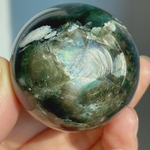 May include: A polished spherical stone featuring deep forest green and dark charcoal tones with iridescent, shimmering inclusions of white and silver mineral deposits that catch the light across its smooth, rounded surface.