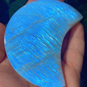 May include: A crescent-shaped, polished gemstone with a vibrant blue hue and iridescent streaks. The stone is held in a hand, showcasing its smooth surface and natural patterns. The gemstone's color shifts with light, creating a captivating display.