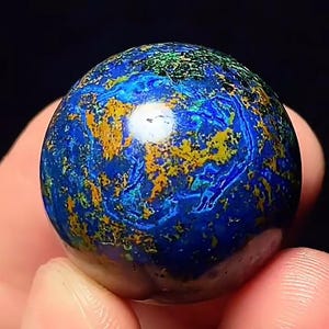 May include: A polished spherical stone featuring a vibrant mix of deep royal blue, bright turquoise, and metallic gold mineral inclusions. The surface displays a swirling, marbled pattern with a high-gloss finish that reflects light.