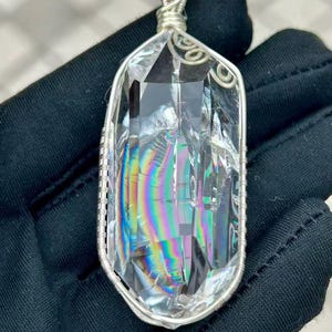 May include: A clear, elongated crystal pendant featuring internal rainbow-colored light refractions. The crystal is encased in a silver-colored wire-wrapped frame with a decorative coiled bail at the top, showcasing intricate metalwork details.