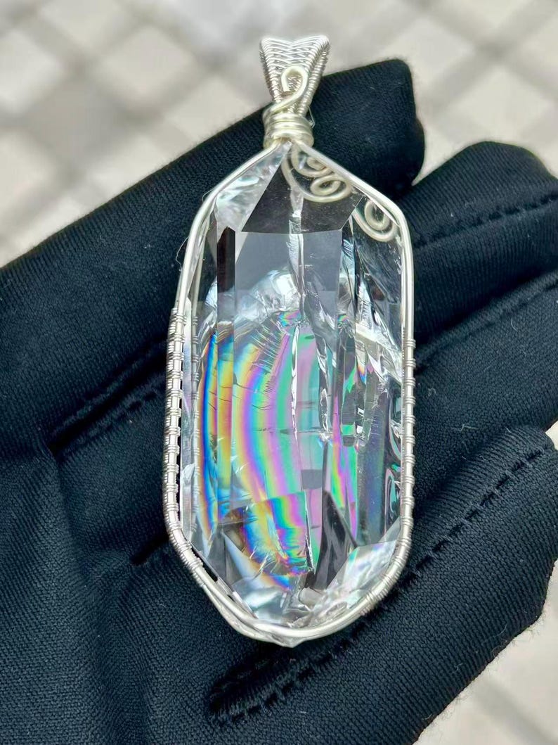 May include: A clear, elongated crystal pendant featuring internal rainbow-colored light refractions. The crystal is encased in a silver-colored wire-wrapped frame with a decorative coiled bail at the top, showcasing intricate metalwork details.