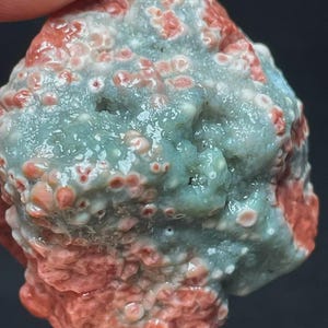 May include: Close-up of a natural stone with a textured surface. The stone features a mix of colors, including red, pink, and a muted blue-green. The surface has small, rounded protrusions.