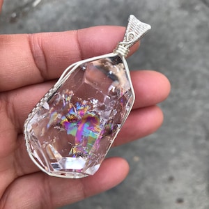 May include: A clear, faceted crystal pendant encased in a silver-colored wire wrap. The crystal features internal fractures that display a vibrant, iridescent rainbow sheen. The wire is intricately coiled around the edges and forms a top loop.