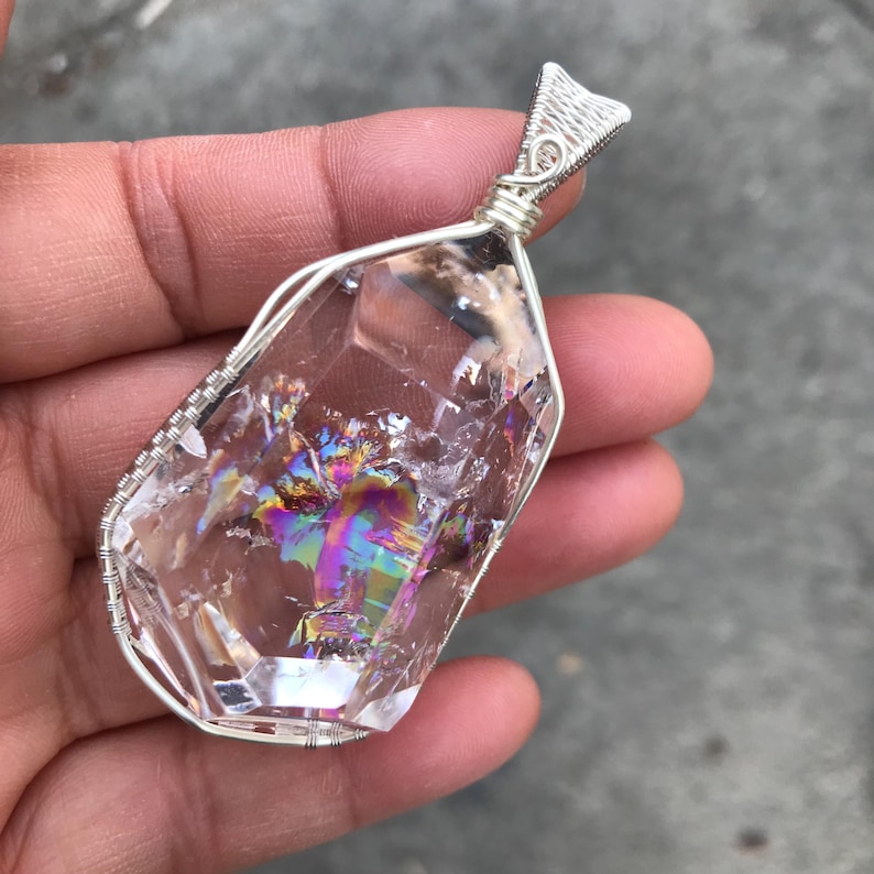 May include: A clear, faceted crystal pendant encased in a silver-colored wire wrap. The crystal features internal fractures that display a vibrant, iridescent rainbow sheen. The wire is intricately coiled around the edges and forms a top loop.