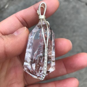 May include: A clear, faceted quartz crystal pendant encased in a silver-colored wire wrap. The wire is coiled tightly around the base and sides of the stone, extending upward to form a secure loop at the top for hanging on a necklace chain.