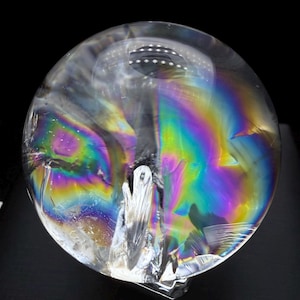 May include: A spherical glass paperweight featuring a clear, transparent body with internal fractures that create vibrant, iridescent rainbow light refractions. The surface is smooth and rounded, displaying a spectrum of colors throughout.