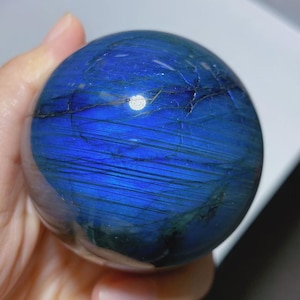 May include: A polished labradorite sphere displaying a vibrant, iridescent blue flash across its surface. The stone features natural dark inclusions and fine linear striations that run horizontally across the rounded, smooth, and reflective mineral form.