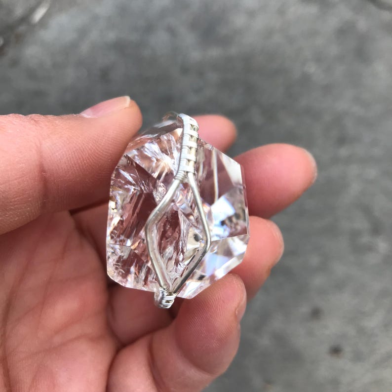 May include: A clear, faceted, irregular-shaped crystal pendant wrapped in thin silver-colored wire. The wire forms a diamond-shaped cage around the center of the stone, with a small loop at the top for hanging. The crystal has multiple sharp edges.