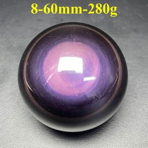 May include: A polished, spherical gemstone with a swirling pattern of purple and black. The text "8-60mm-280g" is visible at the top. The sphere has a glossy finish and reflects light.