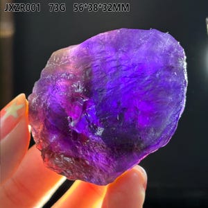 May include: A raw, natural amethyst gemstone. The stone is a deep purple color with lighter areas, and has a rough, textured surface. The dimensions are 56mm x 38mm x 32mm. The weight is 73 grams. The text "JXZR001" is visible.