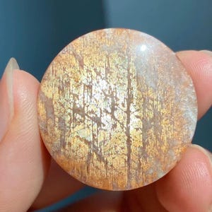 Puede incluir: Un cabujón de piedra de sol circular con una superficie pulida en una paleta de colores naranja y dorado cálido. La piedra preciosa muestra inclusiones metálicas internas que crean un efecto brillante y reflectante en la superficie.