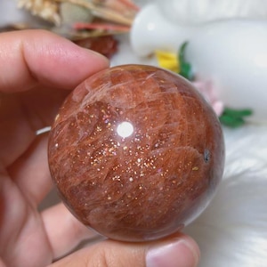 May include: A polished sunstone sphere featuring a deep reddish-orange hue with visible internal fractures and sparkling metallic inclusions that catch the light, creating a shimmering effect across the smooth, rounded surface of the stone.