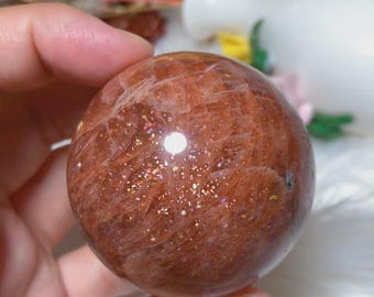 May include: A polished sunstone sphere featuring a deep reddish-orange hue with visible internal fractures and sparkling metallic inclusions that catch the light, creating a shimmering effect across the smooth, rounded surface of the stone.