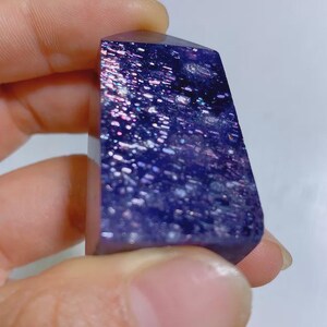 Pu&ograve; includere: Una punta di cristallo lucida di forma rettangolare con un colore di base viola intenso, caratterizzata da numerose pagliuzze metalliche riflettenti incastonate che creano un aspetto scintillante simile a una galassia.