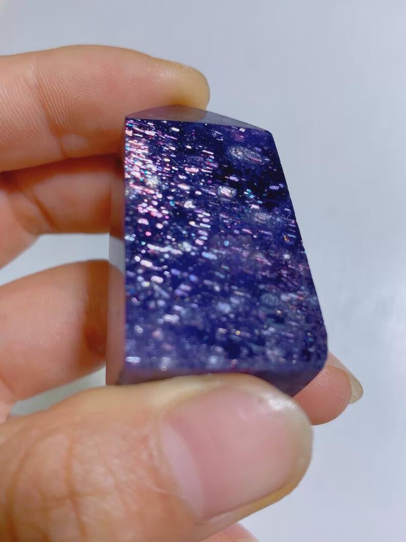 Pu&ograve; includere: Una punta di cristallo lucida di forma rettangolare con un colore di base viola intenso, caratterizzata da numerose pagliuzze metalliche riflettenti incastonate che creano un aspetto scintillante simile a una galassia.