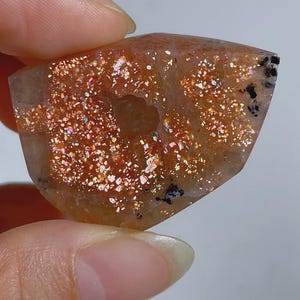 Puede incluir: Una losa de piedra solar pulida con una base translúcida de color melocotón, densas inclusiones metálicas de color cobre brillante y pequeñas motas minerales negras. La piedra tiene una cavidad natural central y muestra un efecto de aventurescencia vibrante y brillante en toda su superficie.