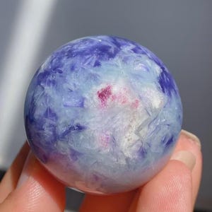 May include: A polished sphere with a mix of blue, purple, and white hues. The crystal ball has a small area of pinkish-red color. The sphere is held in a hand.