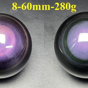 May include: Two polished obsidian spheres. The spheres are black with a purple and pink iridescent center. The text "8-60mm-280g" is in yellow above the spheres. The spheres are approximately 2.36 inches in diameter and weigh 9.88 ounces.