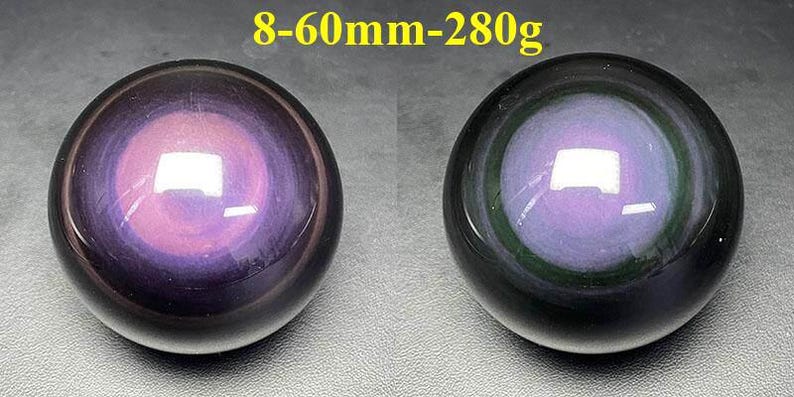 May include: Two polished obsidian spheres. The spheres are black with a purple and pink iridescent center. The text "8-60mm-280g" is in yellow above the spheres. The spheres are approximately 2.36 inches in diameter and weigh 9.88 ounces.