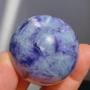 May include: A polished sphere of blue and purple stone. The stone has a crystalline structure with a mix of light blue and deep purple hues. The sphere is held in a hand.