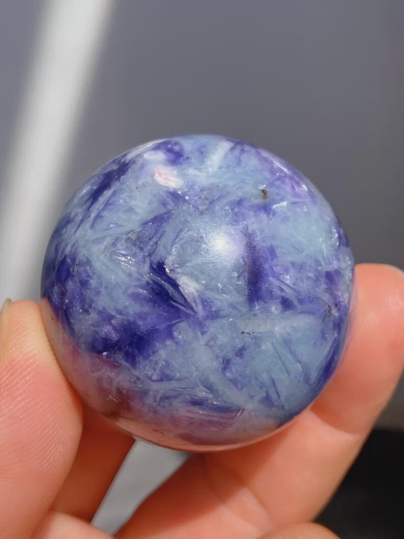 May include: A polished sphere of blue and purple stone. The stone has a crystalline structure with a mix of light blue and deep purple hues. The sphere is held in a hand.
