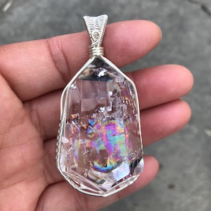 May include: A clear, faceted quartz crystal pendant encased in a silver-colored wire-wrapped frame. The crystal displays internal fractures and a vibrant, multi-colored iridescent sheen throughout its structure, topped with a silver wire bail.