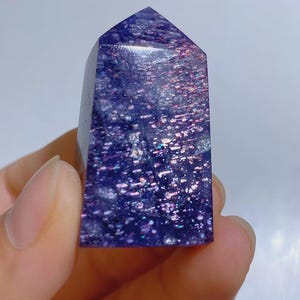 Pu&ograve; includere: Una punta di cristallo lucidata a forma di torre, caratterizzata da un colore di base viola intenso con inclusioni iridescenti simili a glitter che riflettono la luce in tonalit&agrave; rosa, blu e argento sulla sua superficie liscia.
